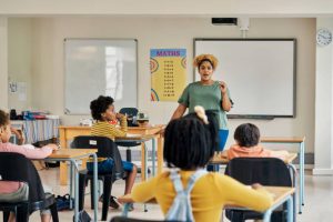 Children classroom, teacher and test writing of kids in a exam lecture hall for knowledge. Learning explanation, student help and school employee in a study lesson with professional black woman